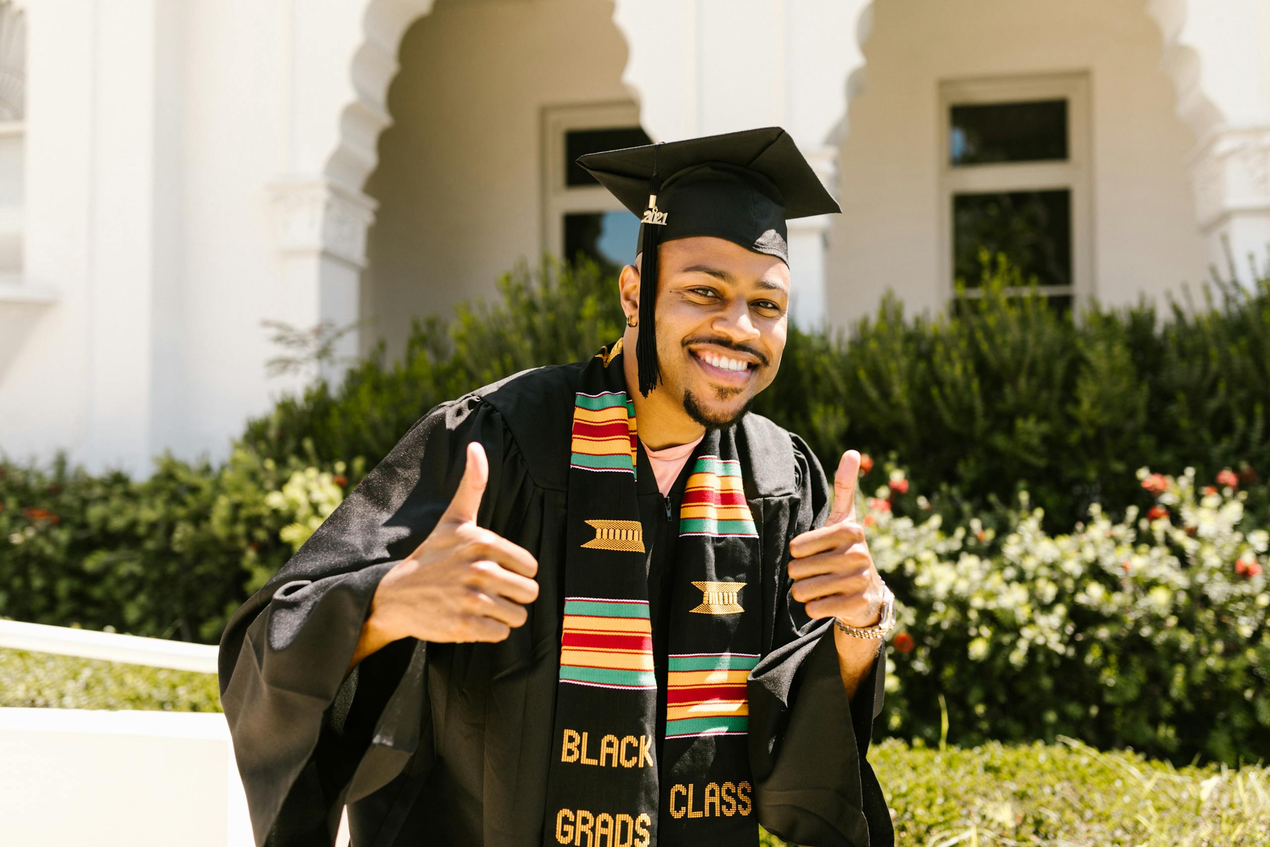 Smiling male graduate outdoors in cap and gown, giving thumbs up. Celebrating achievement.